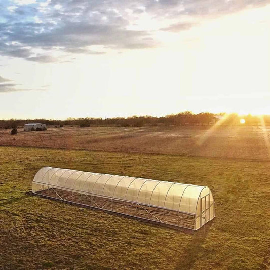 Aerial photograph of a long, transparent greenhouse in a rural field at sunset showcasing golden hour light, a distant farmhouse, and dry grasses.