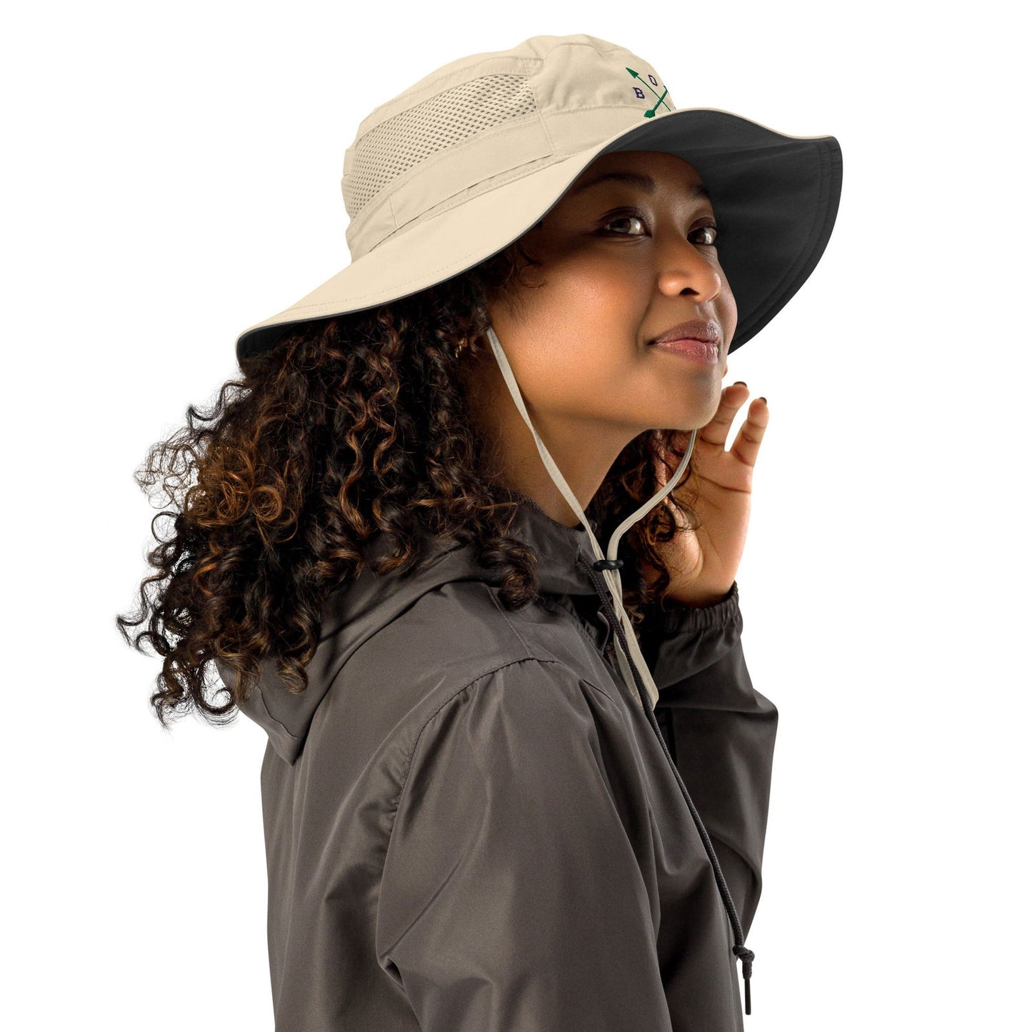 Product shot featuring a woman with curly brown hair wearing a beige and charcoal bucket hat with a logo and a dark gray hooded jacket
