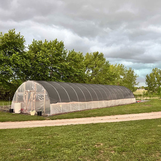 Photograph of a long high tunnel greenhouse in a rural setting featuring lush green trees a dark shade cloth a pale beige ground cover and several black and white plant pots
