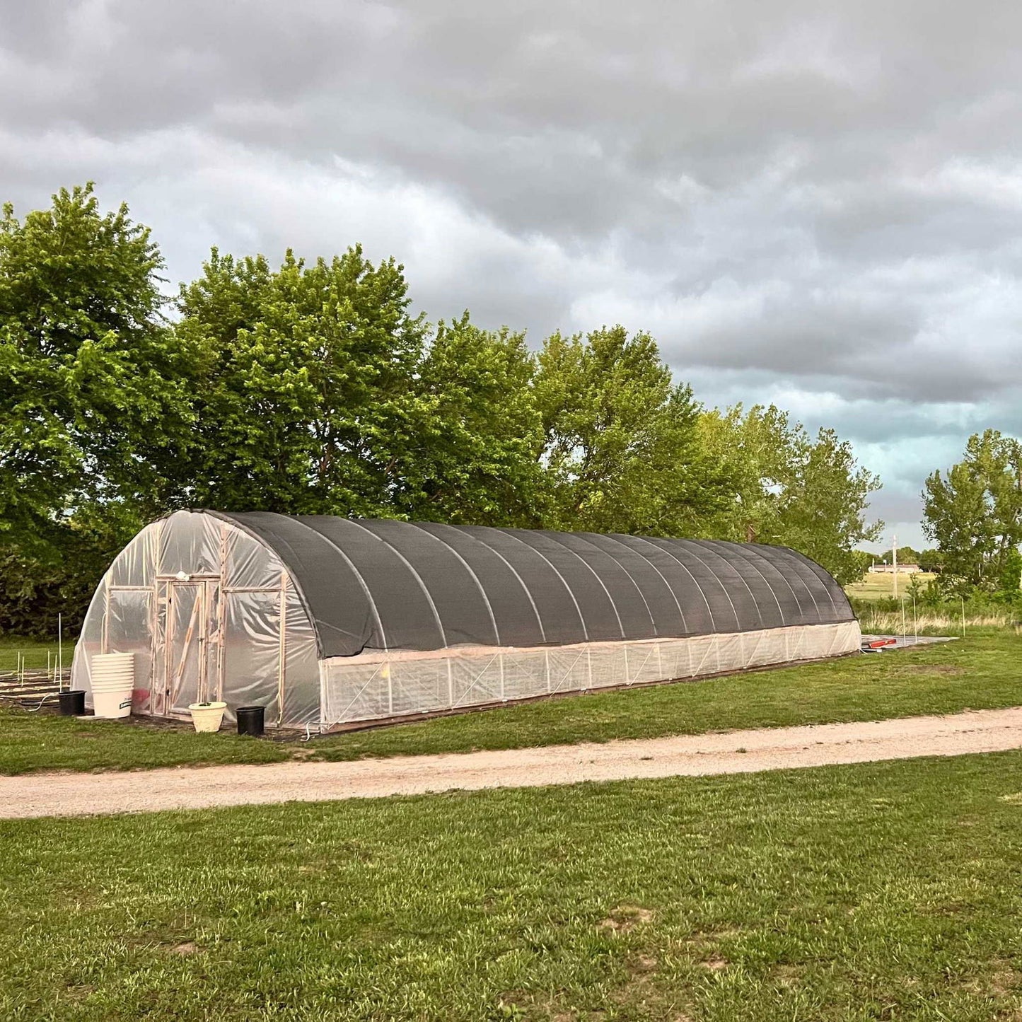 Photograph of a large, high tunnel greenhouse in a rural setting featuring a dark shade cloth, translucent plastic sides, and several potted plants near the entrance under a cloudy sky