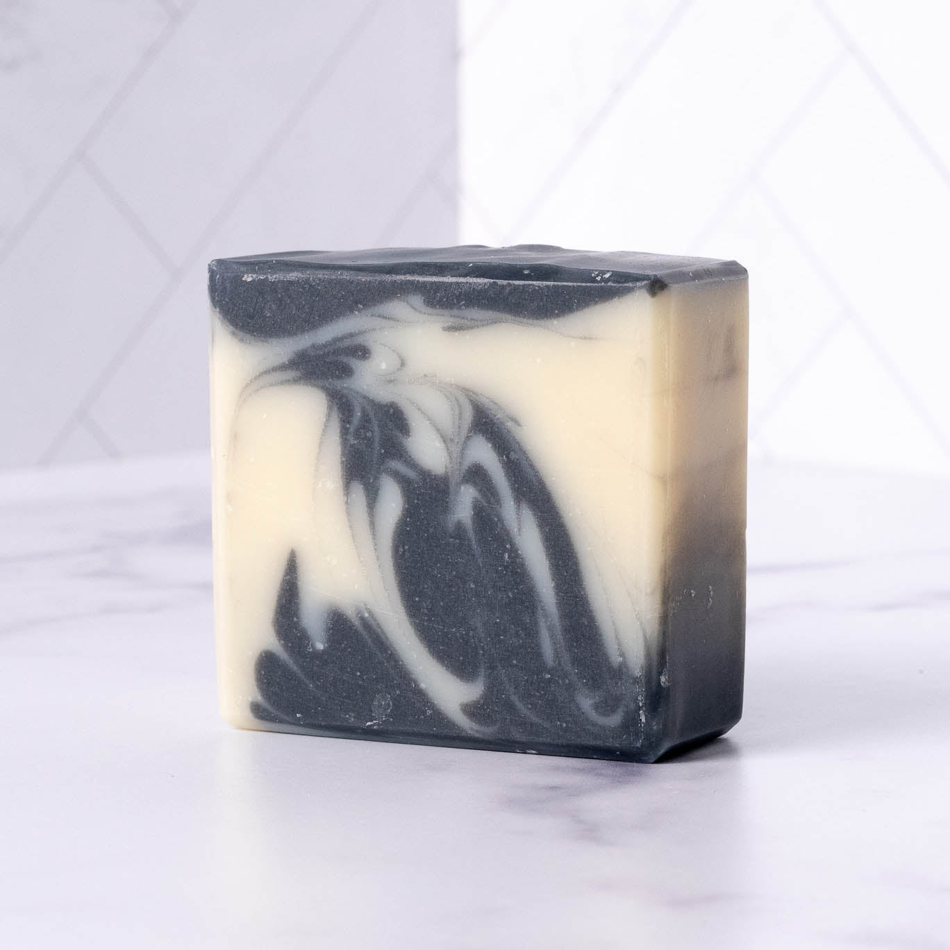 A rectangular Rustic Strength Bar Soap | Detoxifying Charcoal with a marbled black and white pattern rests on a white surface, set against a background of white tiles.