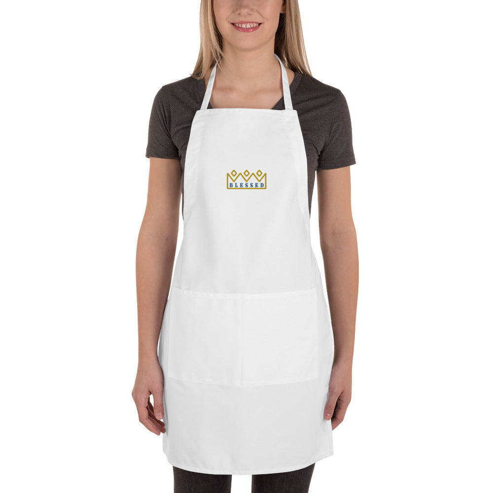 Product shot of a white cotton apron featuring a gold crown and blue Blessed embroidery on a model