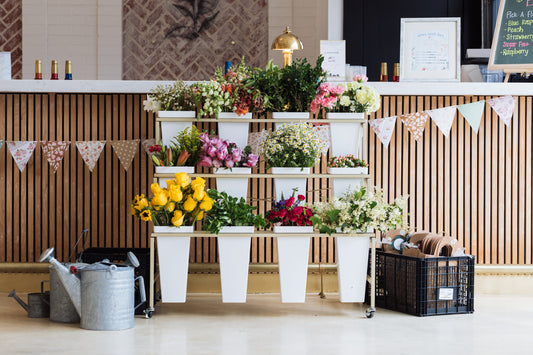 The RENTAL: Flower Display Stand with Buckets from Blessings Grow Meadows Merch & More (Metter, GA pick-up) features white pots for fresh local flowers. Includes buckets, perfect for weddings or events. Watering cans and buntings add a festive touch.