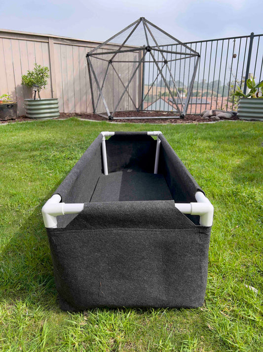 The Fabric Raised Worm Farm by Memes Worms, featuring white PVC pipe supports, sits on backyard grass with a large geometric metal structure and other garden beds in the background.