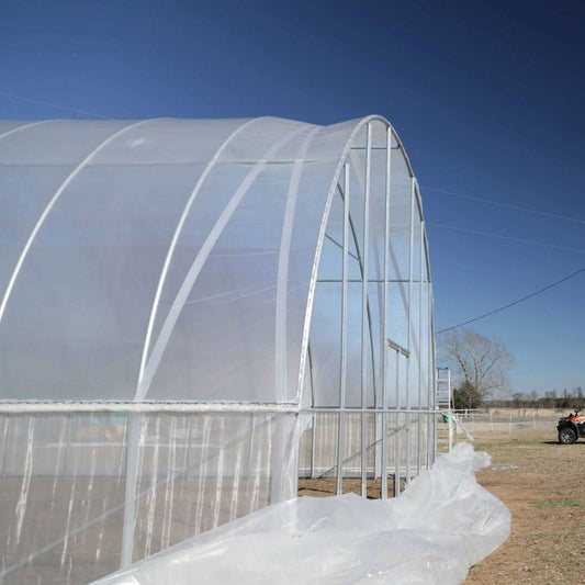 Photograph of a partially constructed high tunnel greenhouse featuring a curved silver frame and translucent white polyethylene sheeting with visible reinforced seams and a small door under a clear blue sky near a fence and ATV