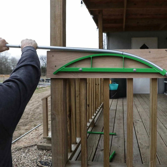 Photo showing a person bending a metal pipe using a bright green pipe bender on a wooden porch with visible wooden railings and a teal bucket in the background