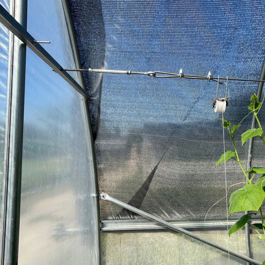 Inside a hoop house, sunlight streams through semi-transparent plastic walls and mesh shade. Metal support bars, including the Bootstrap Farmer Horizontal Bar Kit, span the frame as a vine climbs a string on the right.