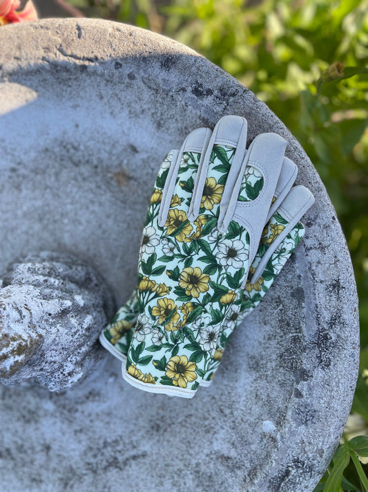 A pair of The Celtic Farm Womens Floral Gardening and Project Gloves The Betsy in green, white, and yellow sits on a weathered stone birdbath, with lush green foliage in the background.