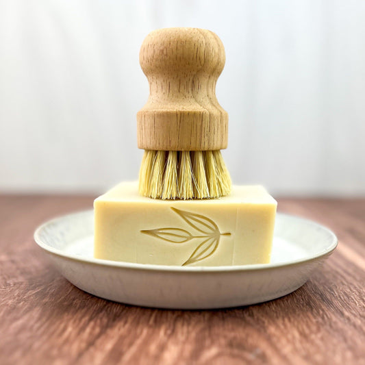 A wooden dish brush stands upright on a rectangular bar of MamaSuds Solid Dish Soap featuring a leaf design, all arranged on a white plate atop a wooden surface.