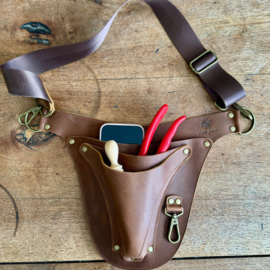 The Celtic Farms Ultimate Leather Garden Tool Belt & Holder—extra rivet reinforced, master gardener designed—securely holds your phone, wooden dibber, and two red-handled pliers for essential tool organization on any surface.