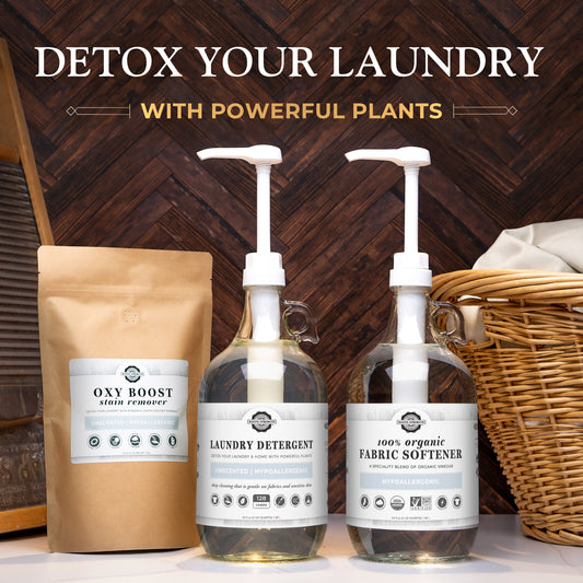 Three eco-friendly Rustic Strength laundry products—a brown pouch of Oxy Boost stain remover, a glass bottle of Laundry Detergent, and a glass bottle of Organic Fabric Softener—rest on a wooden surface beside a laundry basket.