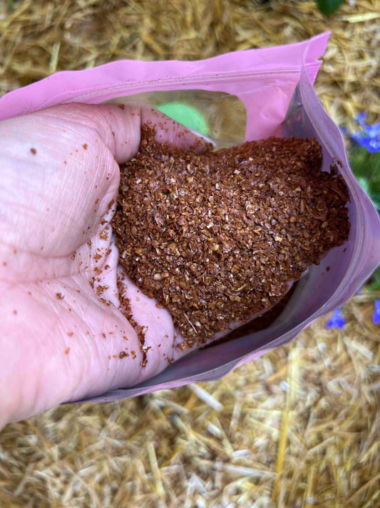 A hand holds coarse, reddish-brown Microbial Compost Food from Memes Worms in a pink resealable bag—ideal for feeding worms and enhancing your compost. Straw and greenery appear in the background.