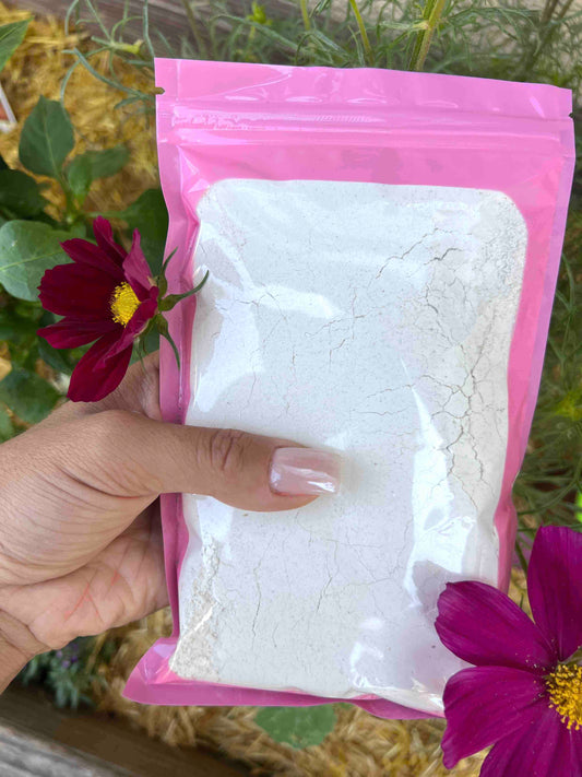 A hand with manicured nails holds a sealed pink plastic bag of Memes Worms Oyster Shell Flour, known for its high calcium content. Purple flowers and green plants appear in the background.