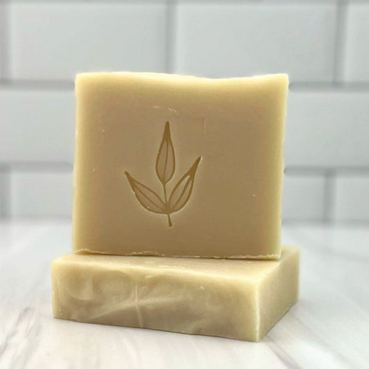 Two beige MamaSuds Scented Castile Bar Soaps are stacked on a white surface with a white tiled background. The top bar has an engraved minimalist leaf design on its front.