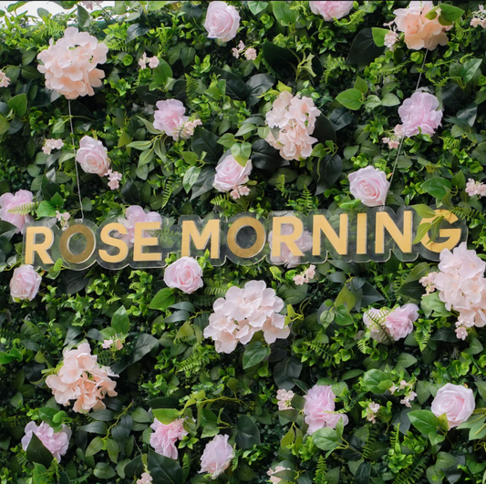 Artificial floral arrangement featuring the words Rose Morning in gold lettering among pale pink roses hydrangeas and lush green foliage.