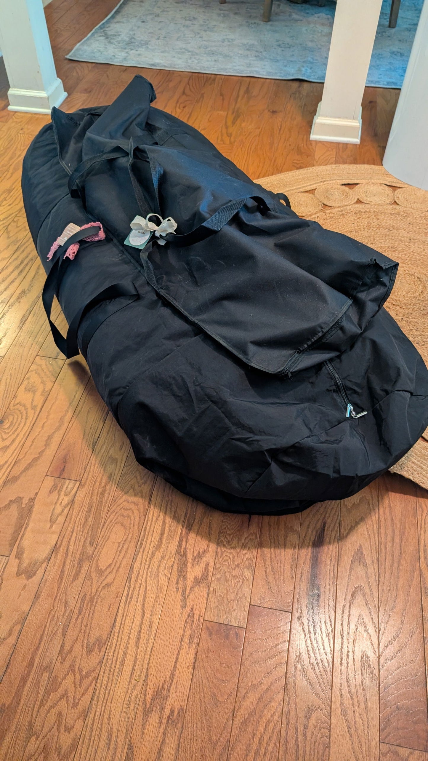 Black fabric storage bag on hardwood floor near white trim and jute rug showing pink ribbon and white tag details