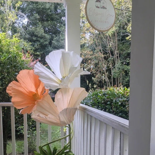 Paper flowers by an unknown artist on a porch overlooking a lush green yard with a Blessings Grow Meadows sign and a white picket fence showcasing peach apricot and ivory blooms