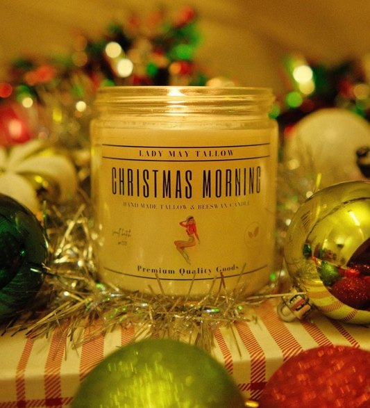 A Lady May Tallow Seasonal Tallow & Beeswax Candle 16oz glows among festive decorations—shiny tinsel and ornaments—creating a warm, all-natural holiday atmosphere with its clean-burning, grass-fed formula.
