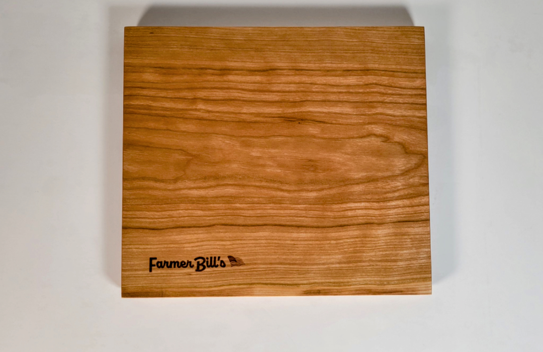 Cherrywood cutting board by Farmer Bill's Provisions, made and sourced in the USA, showing a smooth, polished surface with wood grain details.