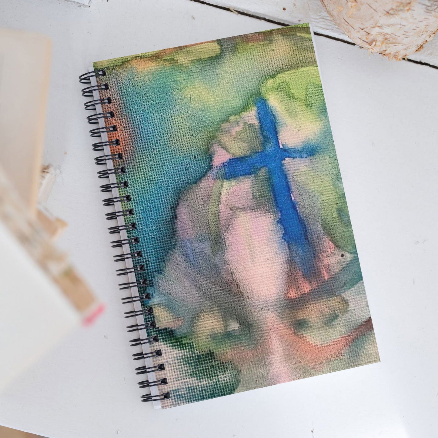 Spiral bound notebook featuring an abstract watercolor painting of a cross with teal, pink, and blue hues on a textured canvas