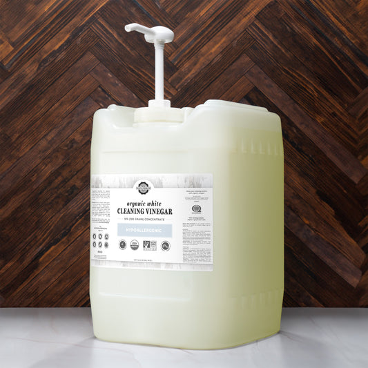 A large Rustic Strength Organic White Cleaning Vinegar Concentrate (10%) container with a pump sits on a white countertop against a dark, herringbone-patterned wood wall.