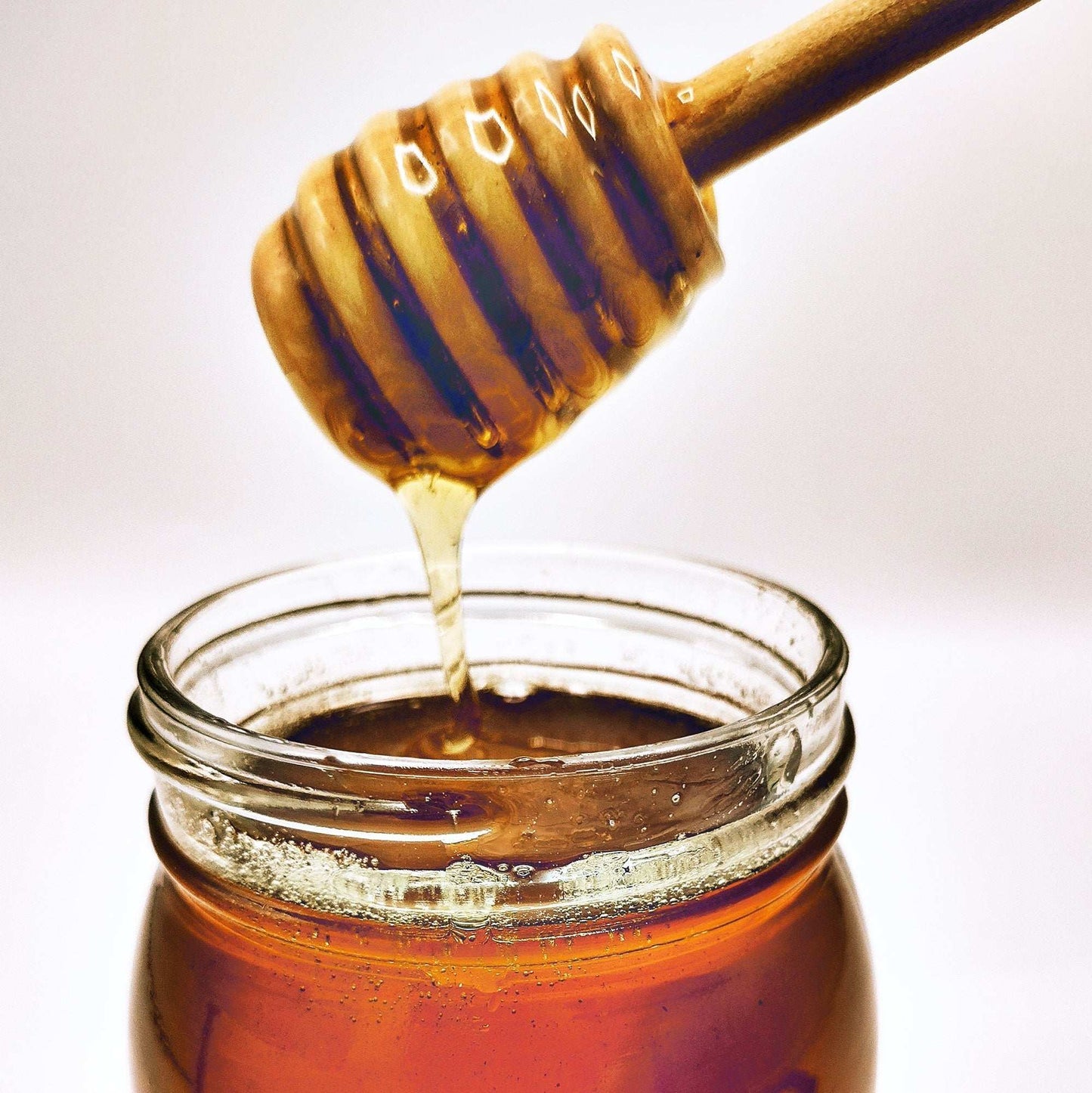A wooden honey dipper drips golden Weeks Raw Gold Reserve Honey, Special Edition—Unfiltered Natural Honey from Georgia by Weeks Honey Farm, into a glass jar against a plain white background.