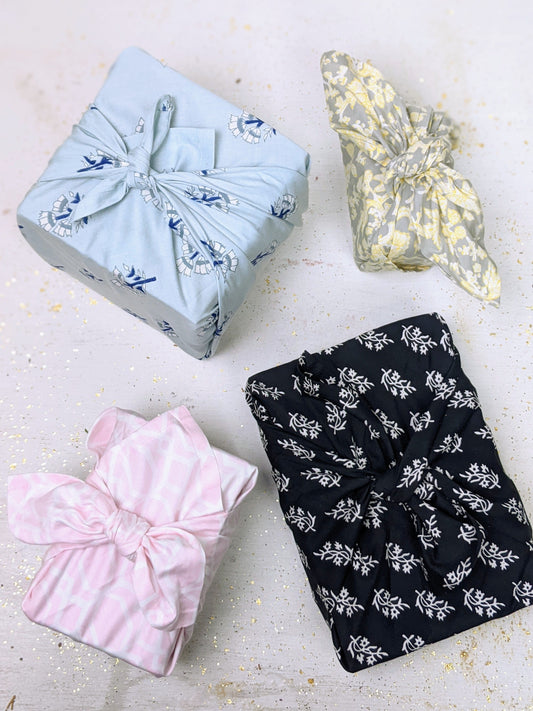 Four rectangular gifts wrapped in patterned fabric, each featuring an Ash & Rose Organic Cotton Bandana—blue with birds, yellow floral, black leaves, and pink geometric—topped with large bows and set on a glitter-sprinkled surface.