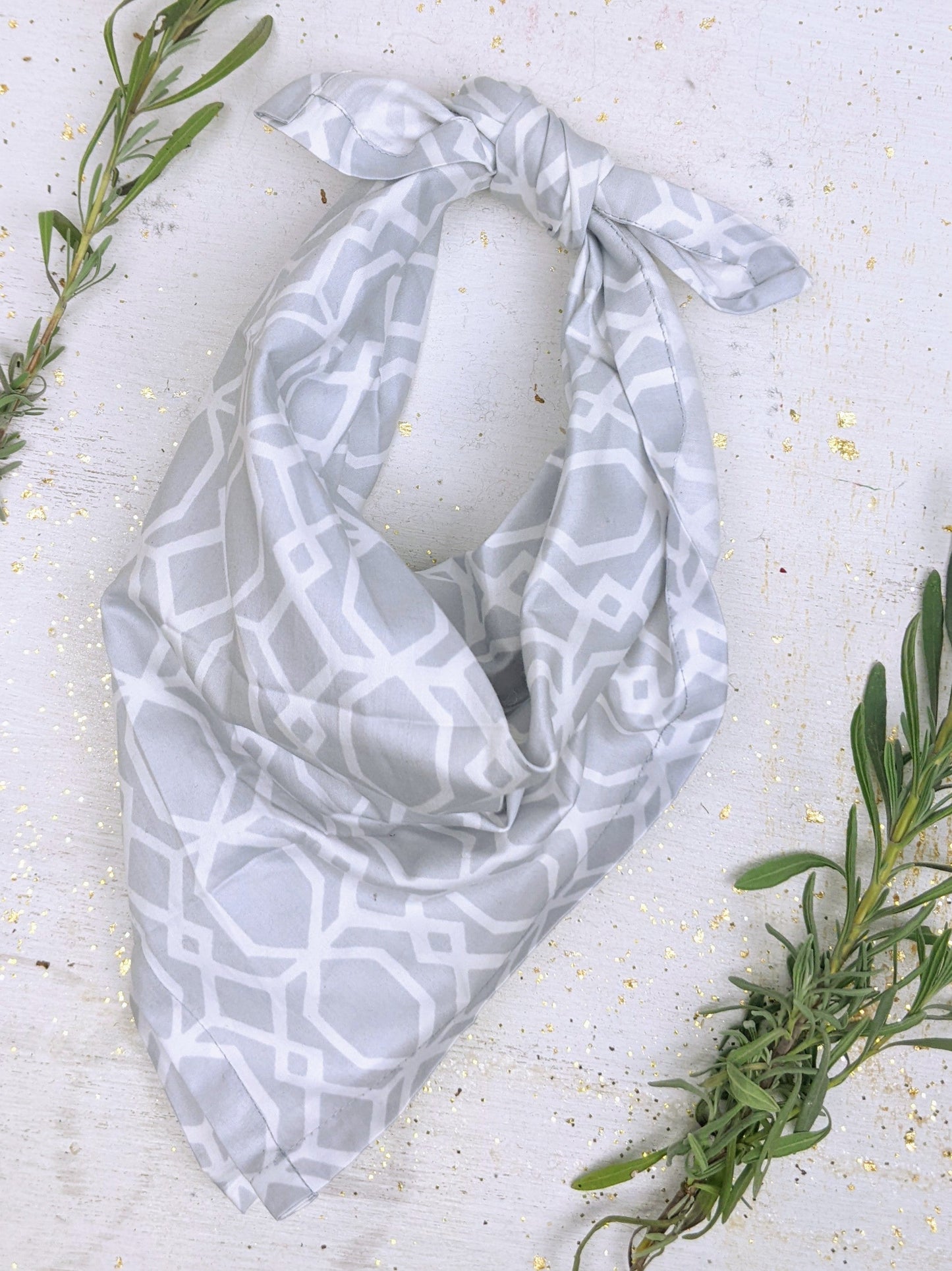 The Ash & Rose Organic Cotton Bandana in light gray with a white geometric pattern is displayed flat on a white background, accented by green sprigs and gold flecks.