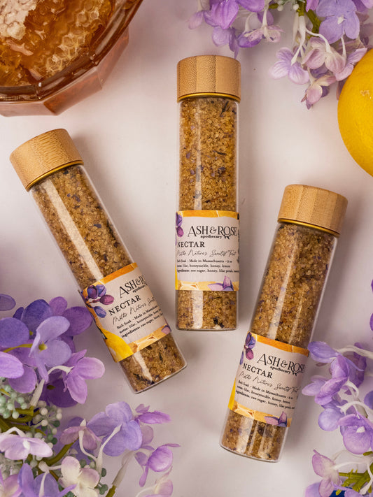 Three glass vials of Ash & Rose NECTAR Bath Soak lie on a white surface, surrounded by lilac petals, purple flowers, a lemon, and a honeycomb dish. The vials feature bamboo lids and floral labels.