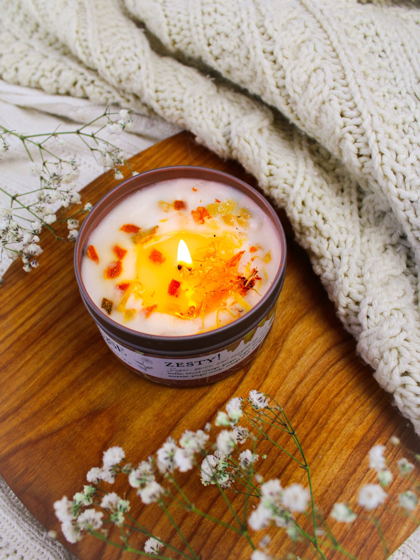 The ZESTY! Orange Ginger Candle by Ash & Rose, handmade and decorated with dried flowers, rests on wood among white baby’s breath and a soft cream knit blanket.