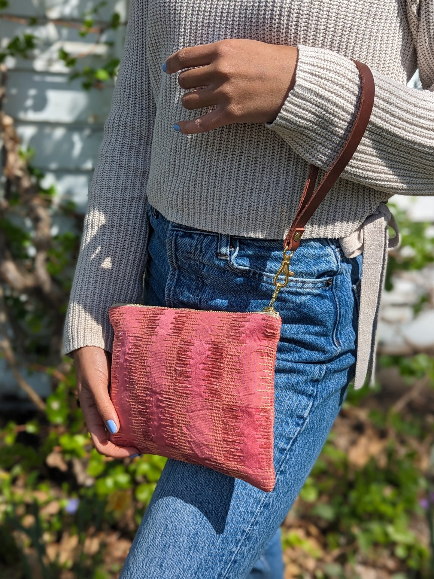 A person in a beige sweater and blue jeans holds the Ash & Rose Shades of Apricot Wristlet Purse, a limited edition pink textured handmade wristlet with a brown strap, while standing outdoors by greenery and a white wall.