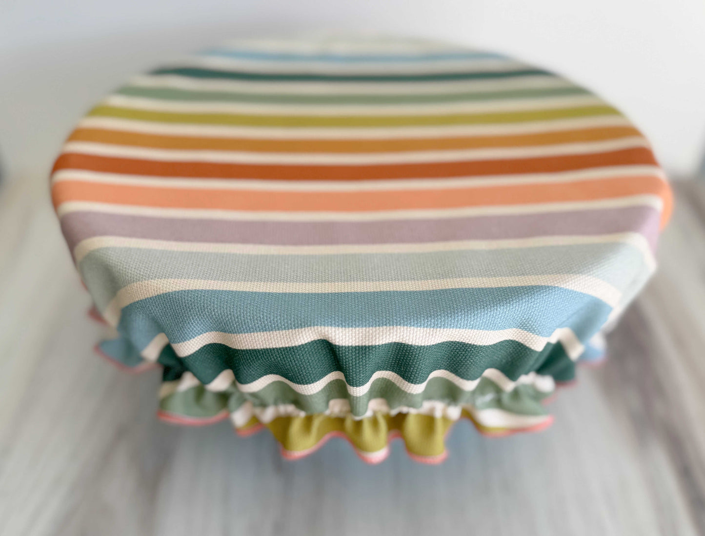 A round bowl topped with the Dot and Army Reusable Bowl Cover—Beach Chair Stripes, featuring horizontal pastels, rests on a light wooden surface.