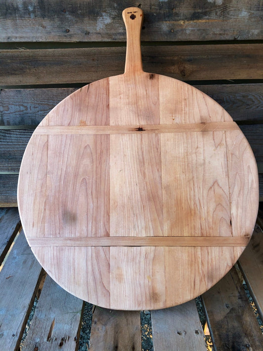 The Celtic Farms Best Charcuterie Board, a vintage-style round maple French breadboard with a handle, rests vertically against a rustic wooden wall on a plank wood surface.