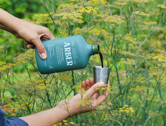 A person pours Arber Organic Bio Protectant, a dark liquid from a green bottle, into a small metal cup while standing outdoors among tall yellow-flowered plants.