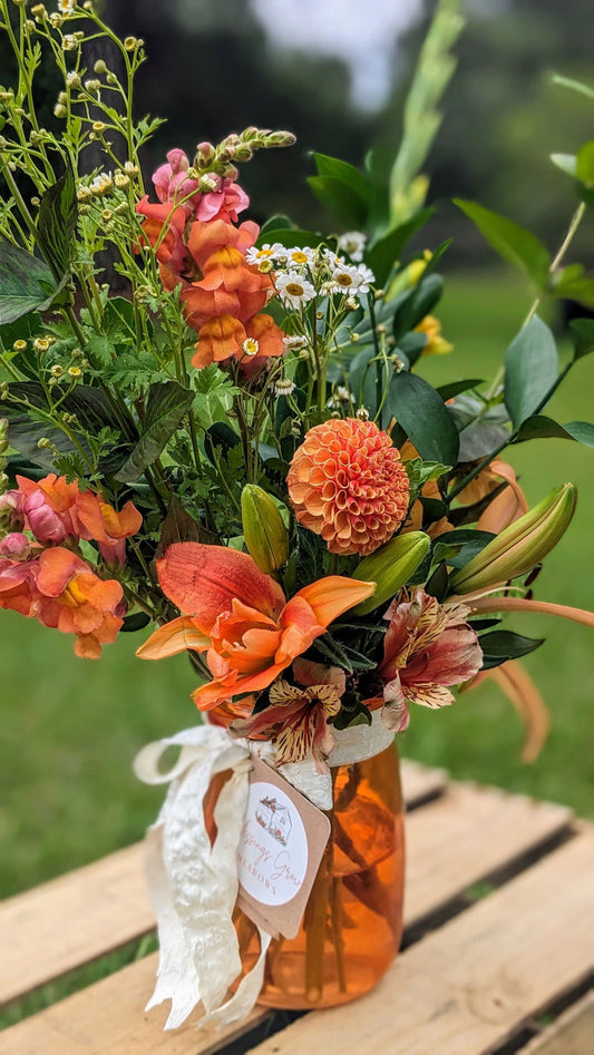 The Blessings Grow Meadows Merch & More Custom Deluxe Arrangement, with orange and yellow dahlias and lilies in an orange glass jar tied with white lace, is displayed on a wooden surface outdoors amid lush greenery.