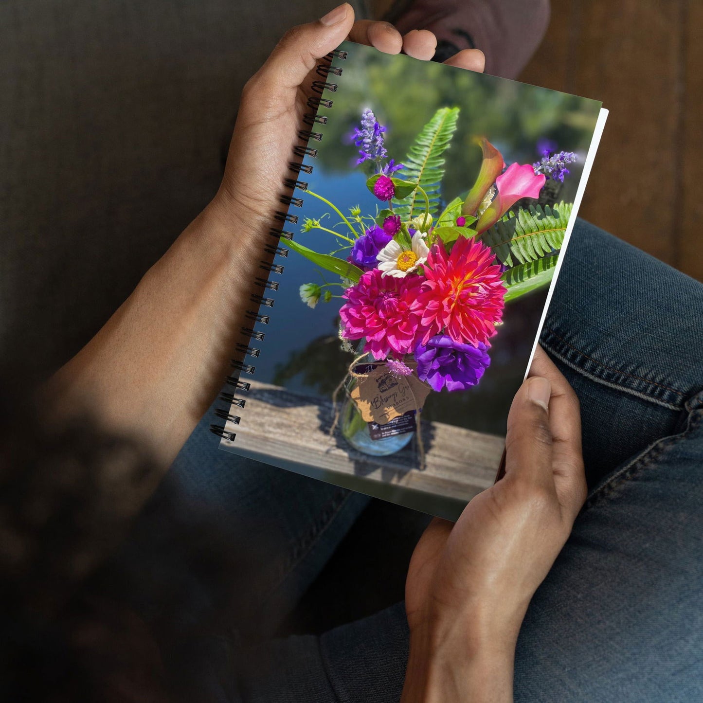 Spiral bound notebook featuring a vibrant floral arrangement of dahlias, calla lilies, and purple lisianthus in a glass jar near a body of water showcasing a fern and wildflowers
