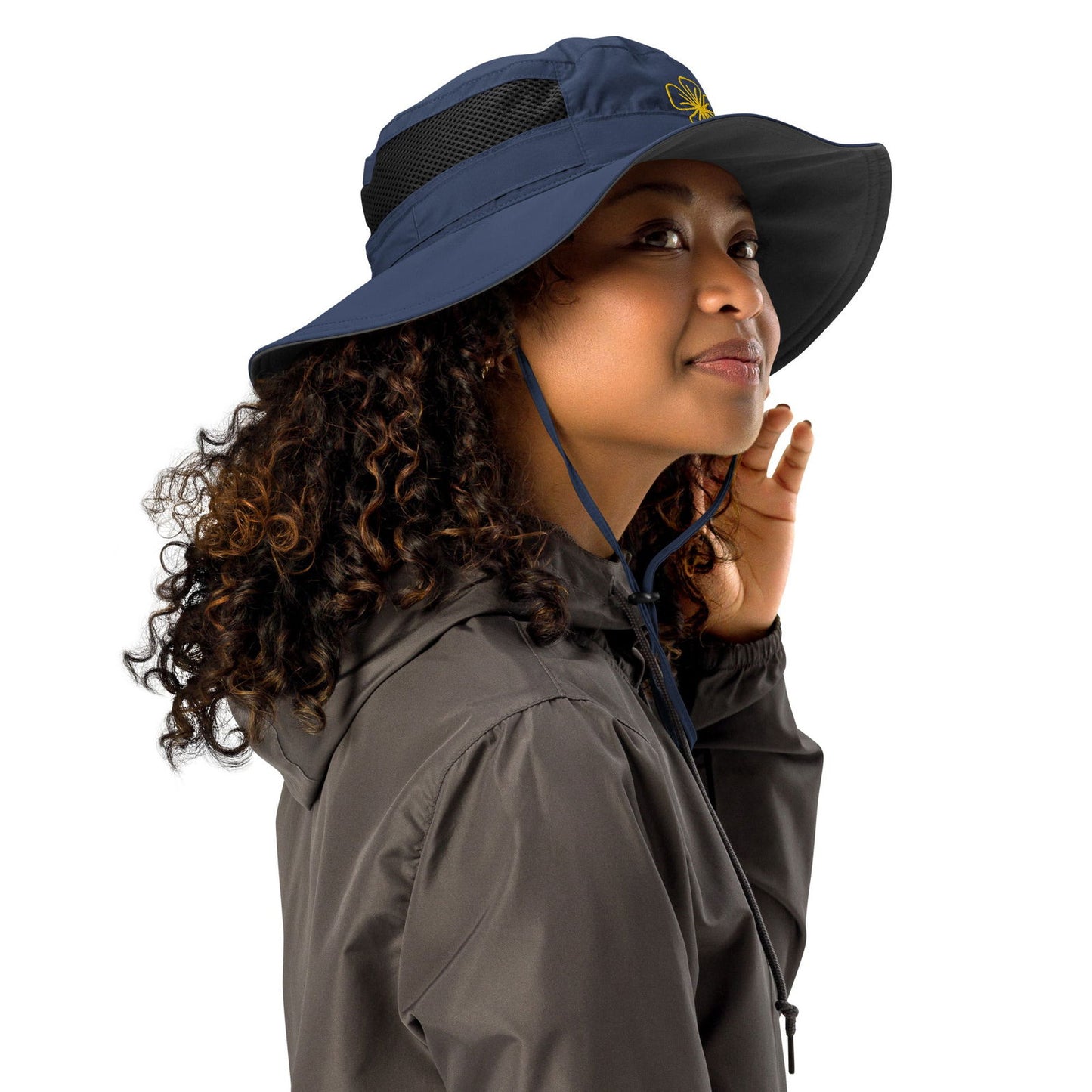 Product image showing a woman with curly brown hair wearing a navy blue sun hat with a mesh insert and gold embroidered detail against a white background.