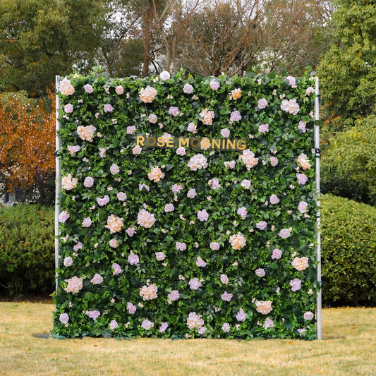 Artificial floral wall installation featuring peach and lavender roses against a backdrop of lush green foliage with the words Rose Morning displayed in gold lettering outdoors near trees