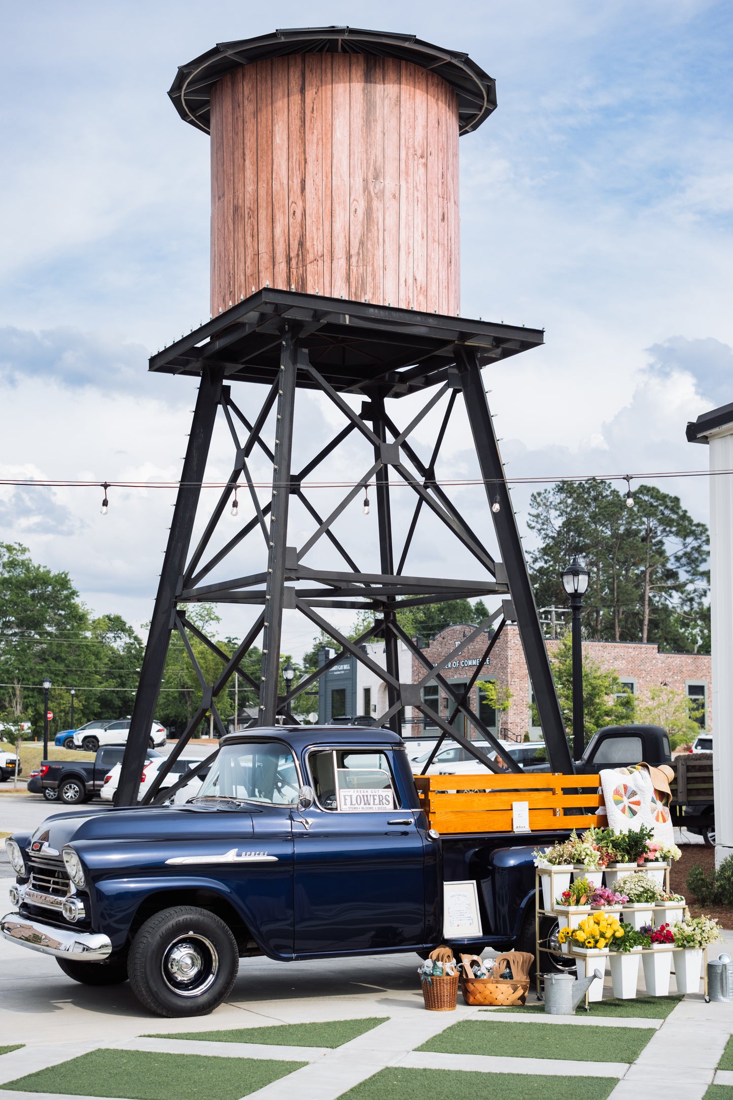 A vintage blue pickup with a wooden flatbed and the Blessings Grow Meadows Merch & More RENTAL: Flower Display Stand with Buckets (Metter, GA pick-up) offers fresh floral arrangements for sale beneath a tall water tower, with trees and buildings in the background.