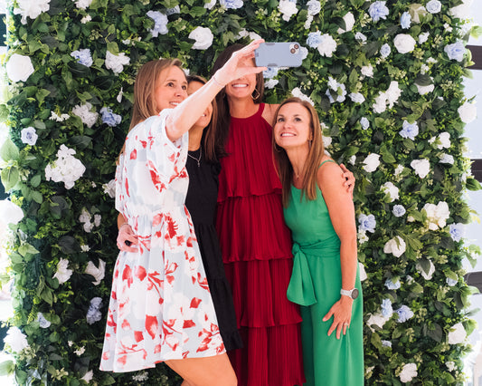 Three women pose for a selfie in front of the RENTAL: Flower Wall Rose + Artificial Flowers Blue & White Curtain Backdrop & Stand 8 x 8 by Blessings Grow Meadows Merch & More, featuring lush greenery and white and blue blooms.