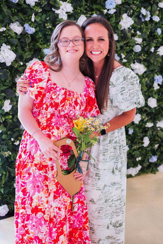 Two women pose in floral dresses—one in bright pinks, the other in soft greens—before the Blessings Grow Meadows RENTAL Flower Wall Rose + Blue & White Curtain Backdrop & Stand (8x8), available for pick-up in Metter, GA. One holds a bouquet and a wooden heart.