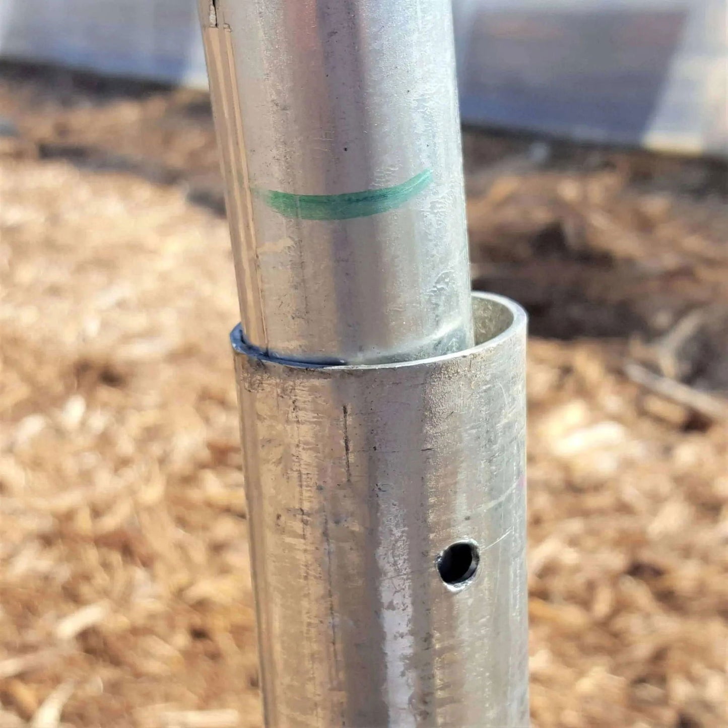 A close-up of Bootstrap Farmer’s Ground Posts - High Tunnel 16 Gauge pipes, one inserted into the other. The outer pipe has a hole, the inner bears a green mark. Brown mulch and a structure are visible in the blurred background.