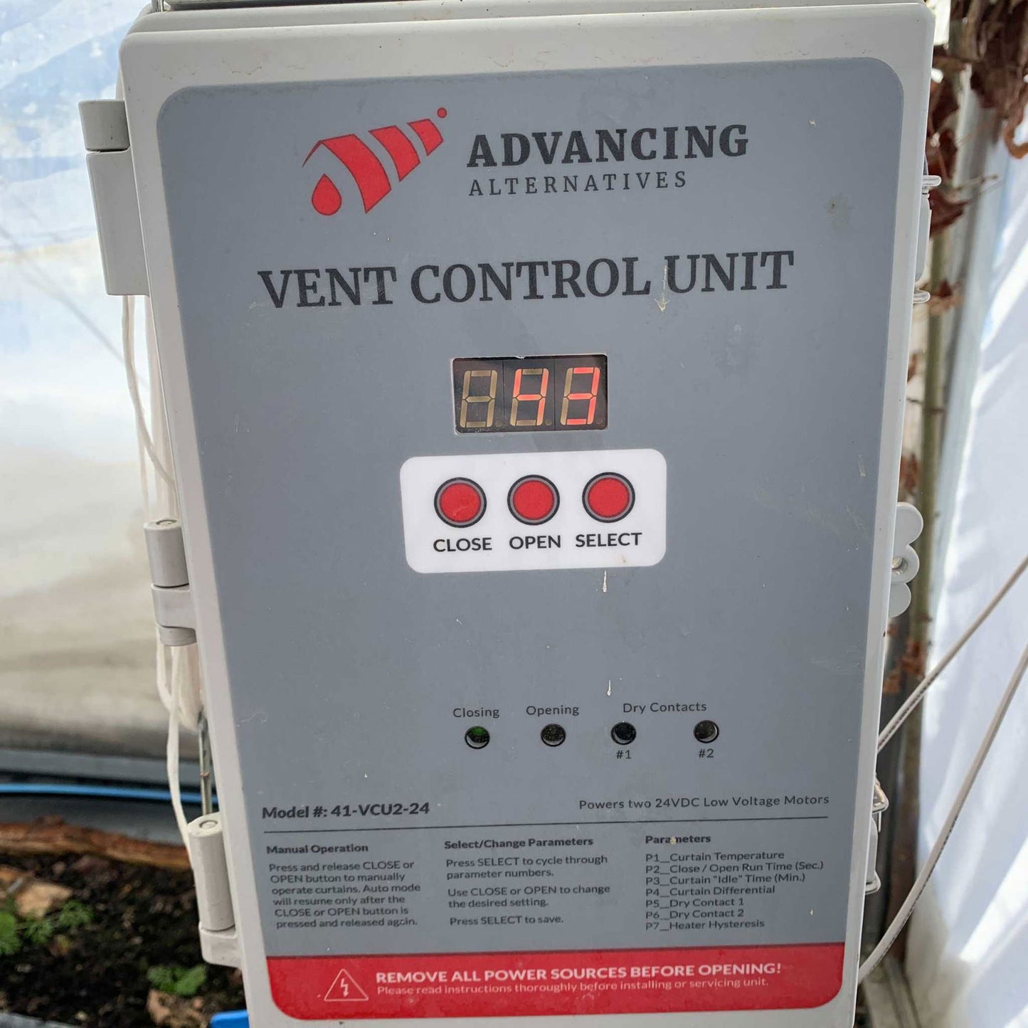A gray Automated Roll-Up Sides Motor by Bootstrap Farmer features a digital display reading 114, buttons for close, open, and select, indicator lights, and a warning label—ideal for automating greenhouse sidewalls.