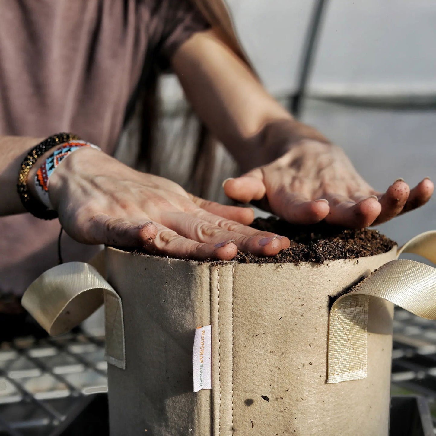Photograph showing a person's hands planting dark soil into a Bootstrap Farmer grow bag with beige fabric and off-white straps outdoors