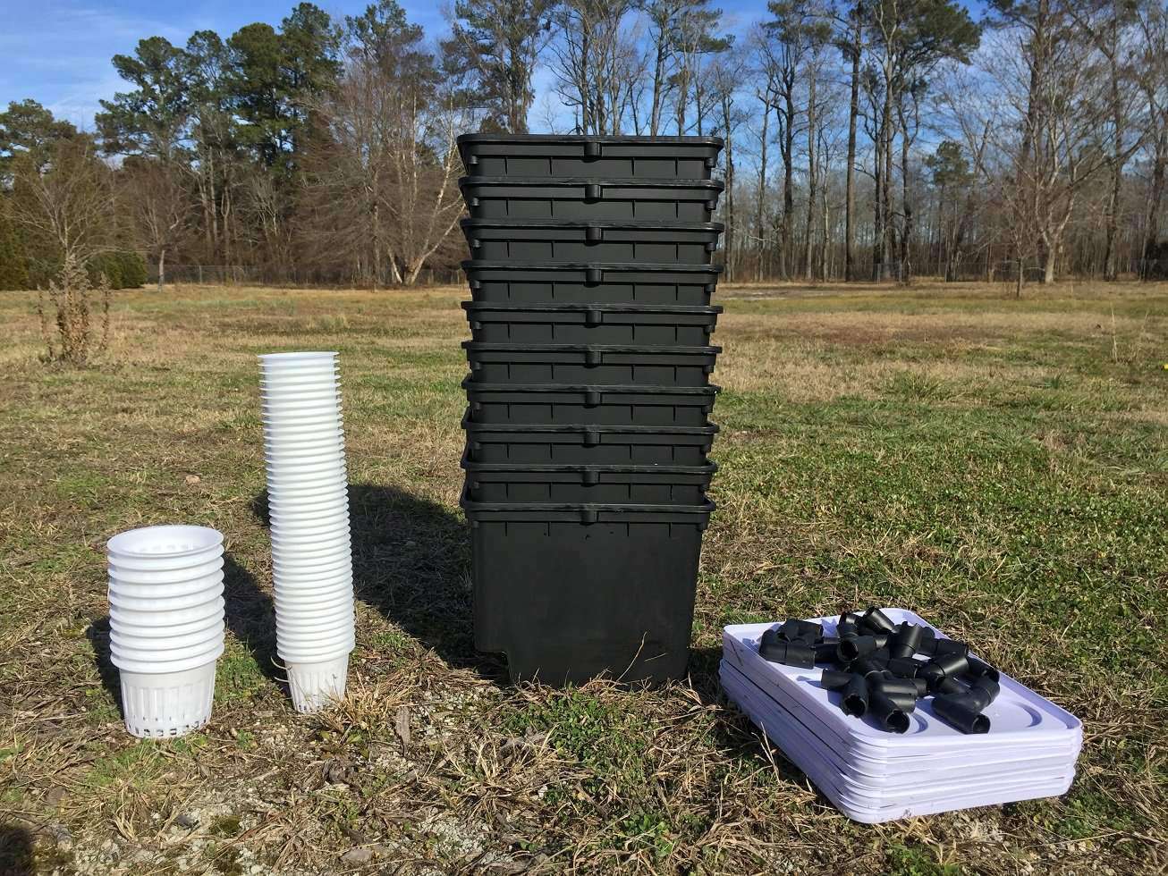 Photograph showing a stack of black plastic containers, stacks of white net pots, and numerous black connectors in a rural field with tall trees in the background.