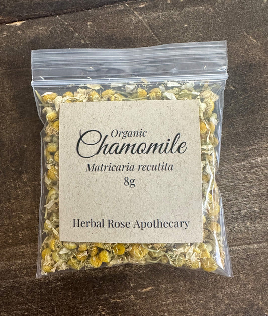 Dried organic chamomile herbs in a clear jar, showcasing small yellow and white chamomile flowers from Herbal Rose Apothecary.