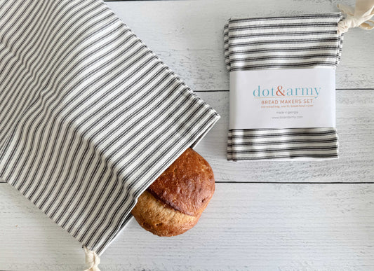A round loaf of homemade bread peeks out of a Dot and Army Grey Ticking Stripe Bread Bag, with a matching folded cloth from the set of two beside it, all displayed on a white wooden surface.