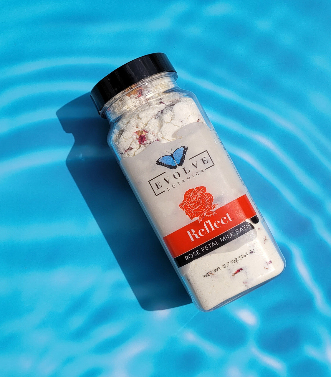 Evolve Botanica Reflect Milk Bath with Rose, creamy powder in a glass jar with pink rose petals and a wooden scoop.