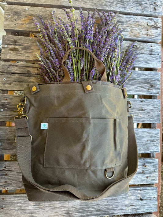 The Celtic Farm Gardeners Multipurpose Shoulder Bag—a brown waxed canvas bag with an outer pocket and adjustable strap—holds fresh purple lavender on a rustic bench, ideal for organized storage of tools, market finds, or harvests.