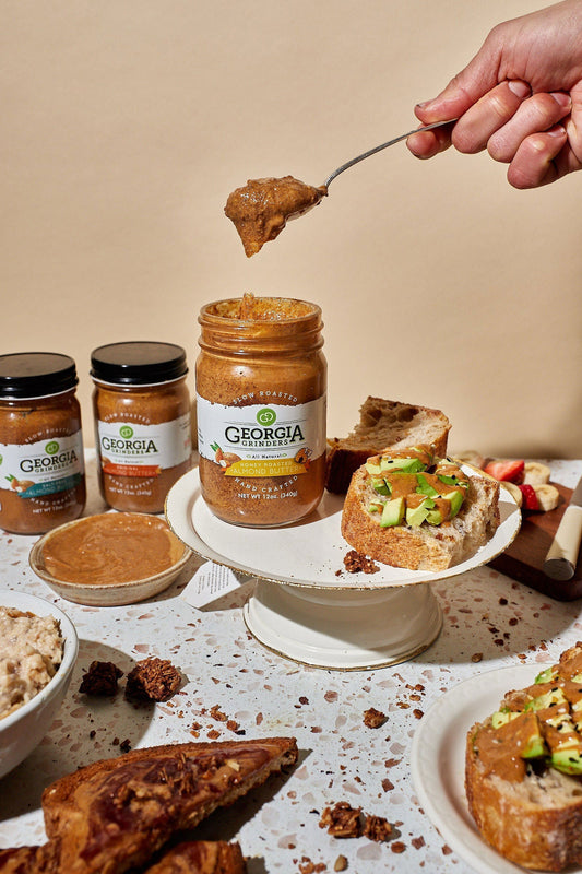 A hand holds a spoonful of Georgia Grinders 64 oz Bulk Tub Honey Roasted Almond Butter over an open jar, surrounded by more jars, sliced bread, and avocado toast—ideal for vegan or keto diets.